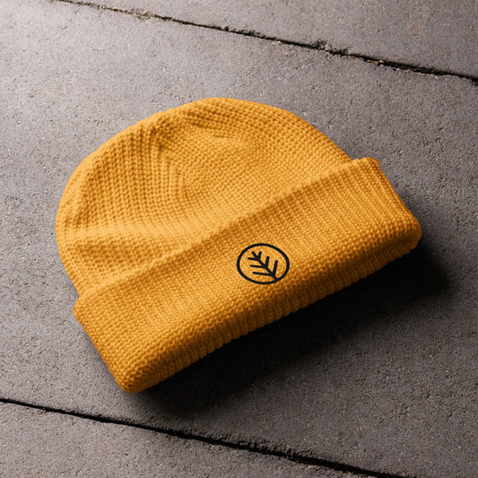 A mustard-yellow knitted beanie lies on a textured concrete surface. The folded brim features a minimalist black embroidered leaf design encased in a circular outline, adding a clean and stylish touch to the cozy accessory.