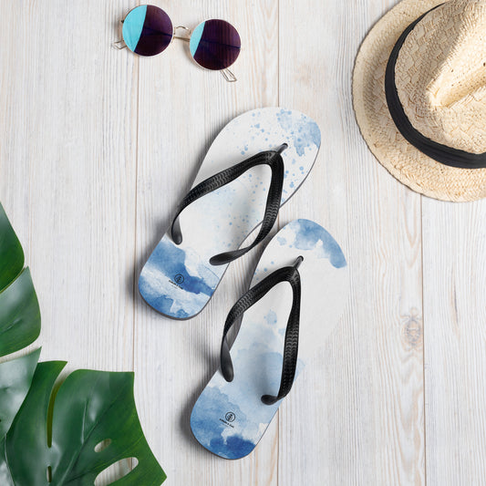 Pair of stylish flip flops with a blue watercolor design, black straps, and a minimalist logo, placed on a light wooden surface next to tropical green leaves, a straw hat, and round sunglasses.