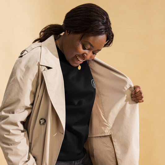 Smiling woman wearing a beige trench coat over a black premium sweatshirt, showcasing a minimal, eco-conscious style by Aurum & Oak. Perfect for sustainable fashion enthusiasts looking for timeless wardrobe essentials.