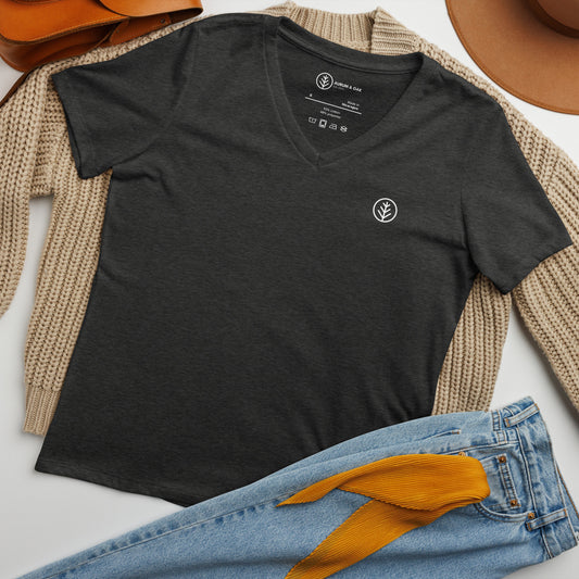 Dark grey V-neck T-shirt from Aurum & Oak with a small leaf logo on the chest, styled with a beige knit sweater, light blue jeans with a yellow scarf in the pocket, a brown leather bag, and a tan hat.