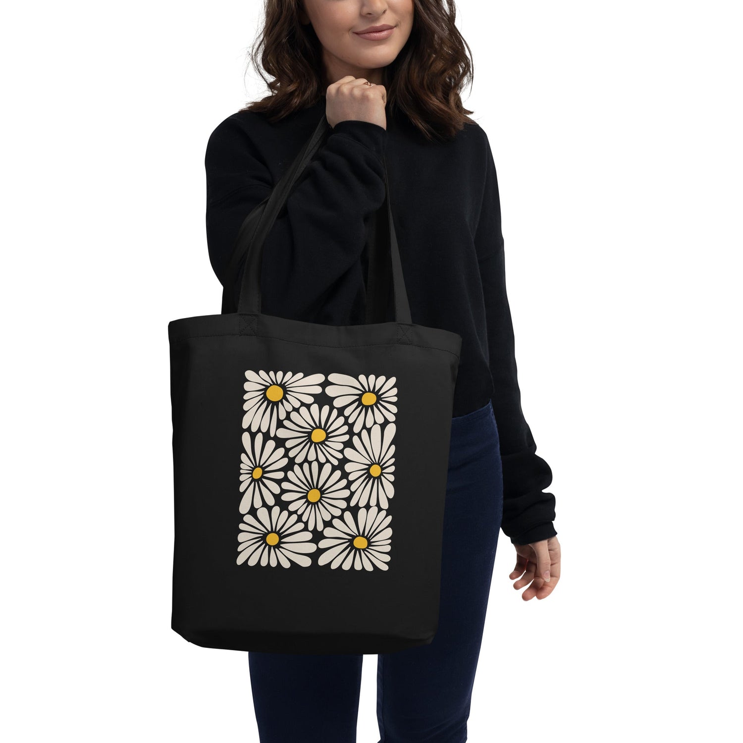 A woman holds a black tote bag featuring a grid of white daisies with yellow centers. Her black sweater and dark pants complement the tote's minimalist, stylish design, creating a chic, modern look that highlights the floral pattern.